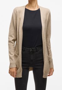Beige cardigan, long sleeves, open front design, ribbed cuffs, pockets on the front, worn over a dark top and black jeans. Smooth texture.