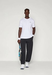 Vyras, vilkintis baltą marškinėlį, juodas sportines kelnes, baltus sportbačius ir apyrankes, laikantis teniso raketę neutraliame studijoje.