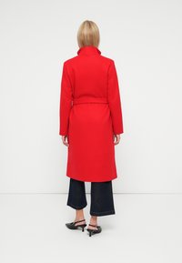 Cappotto lungo rosso con colletto alto, vita cintata e maniche lunghe, abbinato a jeans scuri a gamba larga e scarpe nere con tacco.
