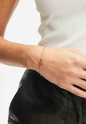 Roze gouden armband van fijne metalen ketting met een minimalistische schuifsluiting. Draagt om een pols tegen een gestructureerde zwarte leren achtergrond.