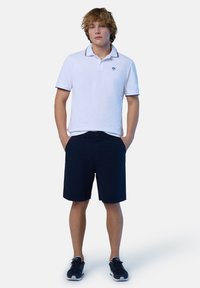 Maglietta polo bianca con bordi blu marina e logo, abbinata a pantaloni corti blu marina. Il materiale sembra morbido, con una vestibilità casual. Sneakers nere completano il look.