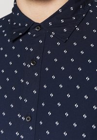 Camisa de botones azul marino con patrones circulares blancos, con cuello y botones negros, hecha de una tela suave.