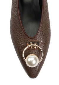 Brązowy, teksturowany but balerina z zaostrzonym czubkiem, czarną wyściółką wewnętrzną i złotym pierścieniem z dużą perłą z przodu.