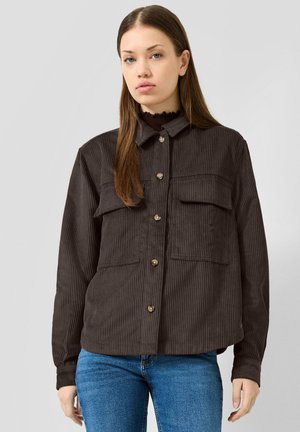 Jeune femme aux longs cheveux portant une veste en velours côtelé marron foncé boutonnée et un jean bleu sur un fond clair uni.