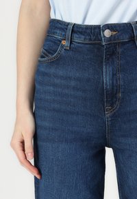Jeans din denim de culoare albastră închisă, cu talie înaltă, cinci buzunare, cusături din cupru și închidere cu buton metalic. Textură netedă și finisaj ușor uzat.