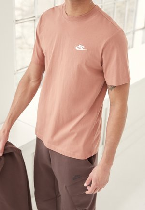 Tričko s krátkým rukávem a kulatým výstřihem v barvě zakouřené růžové, vyrobené z bavlny, s bílým logem Nike na hrudi. Částečně viditelné hnědé kalhoty s kapsou.