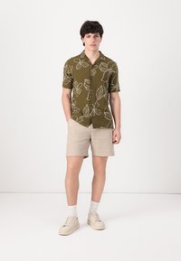 Short-sleeve olive green shirt with white floral pattern, paired with beige shorts and light sneakers, on a model against a white background.