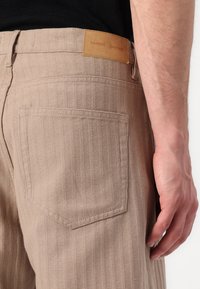 Gestreifte, braune Hose mit Gesäßtaschen, Lederetikett mit der Aufschrift "Samsøe Samsøe" und einem strukturierten Stoff. Sichtbare Nähte.
