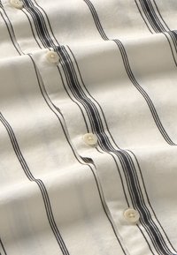 Camisa abotonada de tela color crema con rayas verticales en azul marino oscuro; cuenta con botones blancos y una textura suave y lisa.