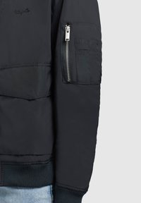 Musta bomber-takki vetoketjullisella hihapussilla, ribbed hihansuilla ja sileällä pinnalla. Ominaisuuksiin kuuluvat brodeeraushaskelmat ja logo-ompeleet.