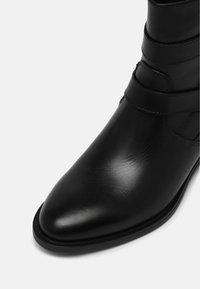 Bottine en cuir noir à bout pointu avec détail de bride, montrant une texture lisse et des coutures sur un fond uni.