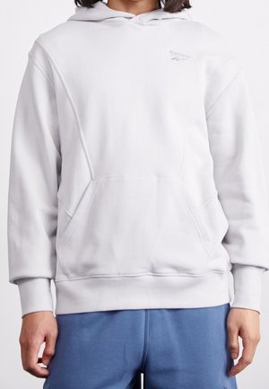 Persoon draagt een effen witte hoodie en blauwe broek, staand tegen een effen witte achtergrond.