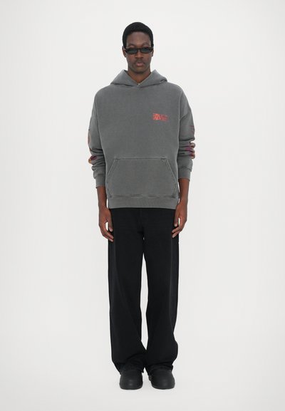 Sweat à capuche gris avec une poche avant, arborant un logo rouge sur la poitrine et des accents colorés sur les manches. Porté avec un pantalon large noir.