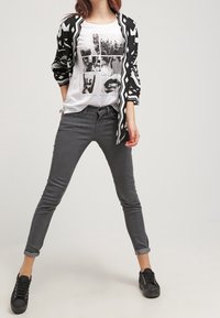 Gestreifter schwarzer und weißer Cardigan über einem weißen Grafik-T-Shirt, kombiniert mit grauen Skinny Jeans und schwarzen Sneakers, ergibt ein lässiges Outfit.