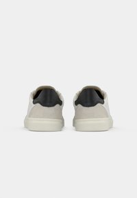 Sneaker in suede bianco e grigio con un accento in pelle nera sul tallone, caratterizzati da una punta arrotondata e suola in gomma, visti da dietro.