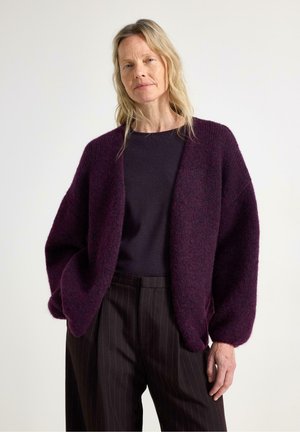 Kvinde med blondt hår iført en mørkelilla cardigan, sort top og sorte stribede bukser mod en ensartet lys baggrund.