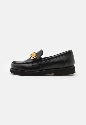 Musta nahkainen loafer-pistokkaalla, paksulla pohjalla ja näkyvällä kultaiseen sävyyn metalliosalla päällisessä, jossa ommeltu moc toe -muotoilu.