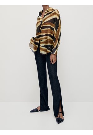 Person iført en løs, tigerstribet skjorte med knapper, mørke, lige bukser med sideslidser og sorte spidse flade sko med ankelstropper.