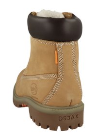 Bota corta de ante color canela con un cuello de cuero marrón, costuras blancas y una suela de goma con un patrón de dibujo resistente. Incluye un detalle en color naranja.