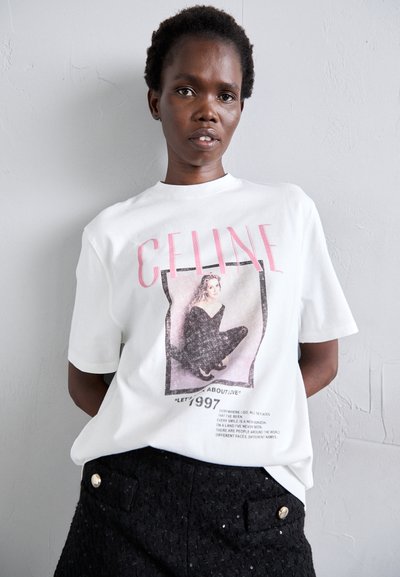 T-shirt en coton blanc avec un motif graphique et un texte rose, porté par un modèle en tenue noire. Le t-shirt a un col rond et des manches courtes.