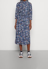 Modré květinové šaty s tříčtvrtečními rukávy, zúženým pasem a knoflíkovým zapínáním vpředu. V kombinaci s černobílými teniskami.
