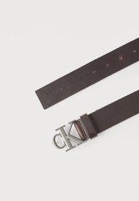 Σκούρο καφέ δερμάτινη ζώνη με ασημί αγκράφα "CK" και πέντε τρύπες ρύθμισης, απεικονίζεται σε λευκό φόντο.