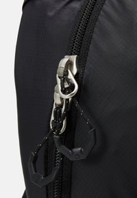Détail d'un sac à dos en tissu noir avec une fermeture éclair en métal et des cordons noirs avec des poignées en plastique. Le matériau a une finition texturée.