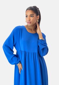 Blaues langärmeliges Kleid mit rundem Ausschnitt, fließendem Oberteil und geraffter Taille. Glatte Textur und entspannte Silhouette, mit goldenen Accessoires.