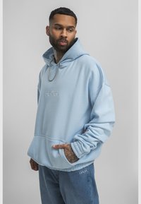 Man with tattoos wearing light blue hoodie and matching jeans stands with one hand in pocket against plain background.