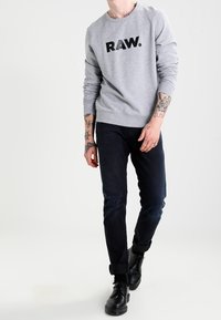 Sudadera gris con el logo "RAW." en negro, combinada con jeans oscuros y botas negras. Presenta tatuajes en los brazos y las mangas enrolladas.