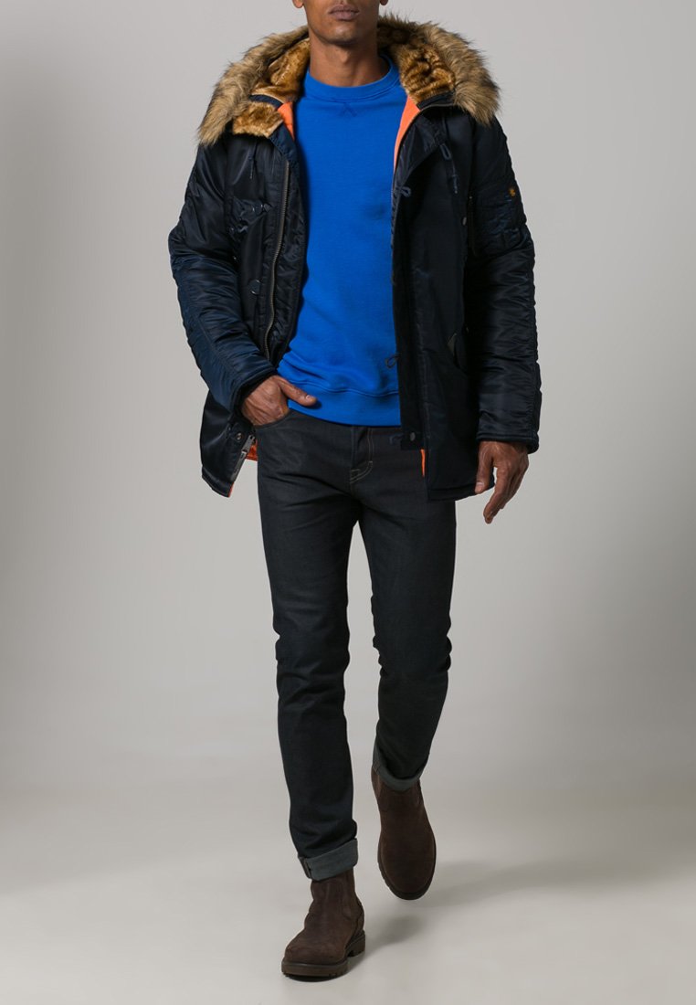 Parka navy con cappuccio in pelliccia sintetica, dettagli arancioni e multiple tasche. Indossato sopra un felpa blu e abbinato a jeans scuri e stivali marroni.