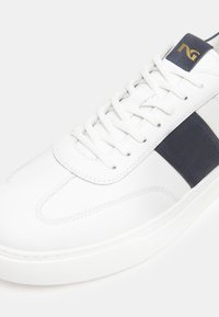 Sneaker bianco con una striscia blu navy, realizzato in pelle liscia. Caratterizzato da lacci piatti e un colletto imbottito. Suola in gomma testurizzata.