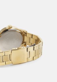Reloj de pulsera de metal dorado con una pulsera articulada, caja redonda y un cierre sencillo. La parte trasera muestra el nombre de la marca y los detalles del modelo grabados.