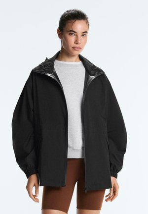 Femeie purtând o jachetă neagră largă cu fermoar peste un hanorac gri deschis și pantaloni scurți de ciclism maro, stând pe un fundal simplu.