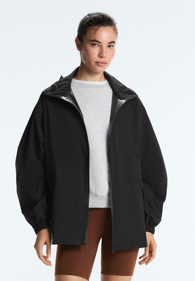 Frau trägt eine lockere schwarze Reißverschlussjacke über einem hellgrauen Sweatshirt und braunen Radhosen, steht vor einem schlichten Hintergrund.
