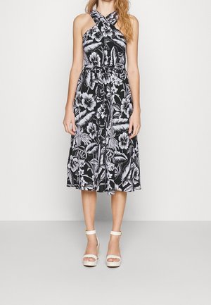 Kobieta w czarno-białej sukience midi w kwiaty z wiązanym w talii dekoltem halter, w połączeniu z białymi sandałami na platformie z paskiem wokół kostki, stojąca na jednolitym tle.