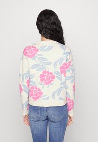 Gestrickter Pullover mit cremefarbenem Grundton, verziert mit pinken Blumenmotiven und blauen Blättern. Das Design wirkt weich und strukturiert, mit lockerer Passform.