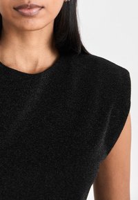 Femme aux cheveux lisses et foncés portant un haut noir texturé sans manches, montrée des épaules aux lèvres sur un fond uni.