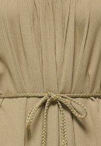 Tissu beige avec des plis verticaux, noué à la taille par une ceinture en corde tressée formant un nœud en boucle.