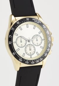 Montre chronographe dorée avec une lunette noire, un cadran argenté et trois sous-cadrans. Équipée d'un bracelet en caoutchouc noir et de grands marqueurs d'heure.