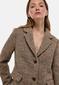 Veste à motifs en chevrons marron avec revers à cran, deux poches avant et trois boutons noirs à l'avant.