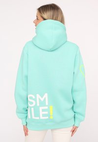Turquoise hoodie made of soft fabric, featuring a large white "SMILE!" text and a small yellow accent heart on the sleeve.