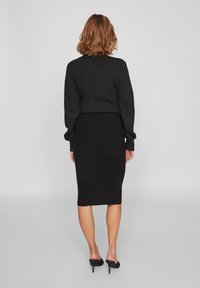 Černý pletený svetr a úzká sukně, ukazující žebrovanou strukturu, přiléhavý střih a design s otevřeným zadním dílem, doplněné černými lodičkami.