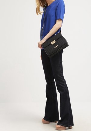 Borsa nera con texture e chiusura in metallo dorato; indossata con una maglietta blu e jeans neri svasati, dotata di tracolla a tracolla.