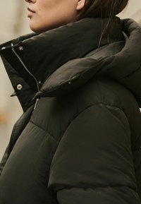 Jachetă puf din negru cu guler înalt, având o textură lucioasă și cusături vizibile, evidențiind detalii de hardware cu nasturi și fermoar.