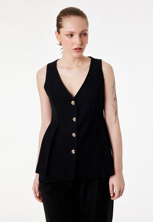 Donna con pelle chiara e tatuaggi indossa un gilet nero senza maniche con bottoni d'oro, guardando leggermente verso destra su uno sfondo bianco.