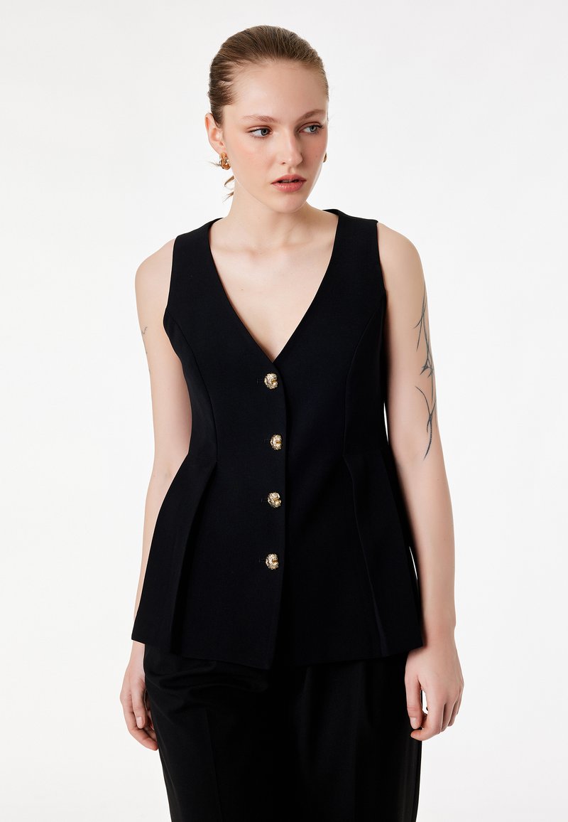 Donna con pelle chiara e tatuaggi indossa un gilet nero senza maniche con bottoni d'oro, guardando leggermente verso destra su uno sfondo bianco.