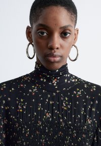 Schwarze Bluse aus Stoff mit hohem Kragen, verziert mit einem Blumenmuster in Rot, Gelb und Weiß. Strukturiertes und gerafftes Design. Silberne Creolen.