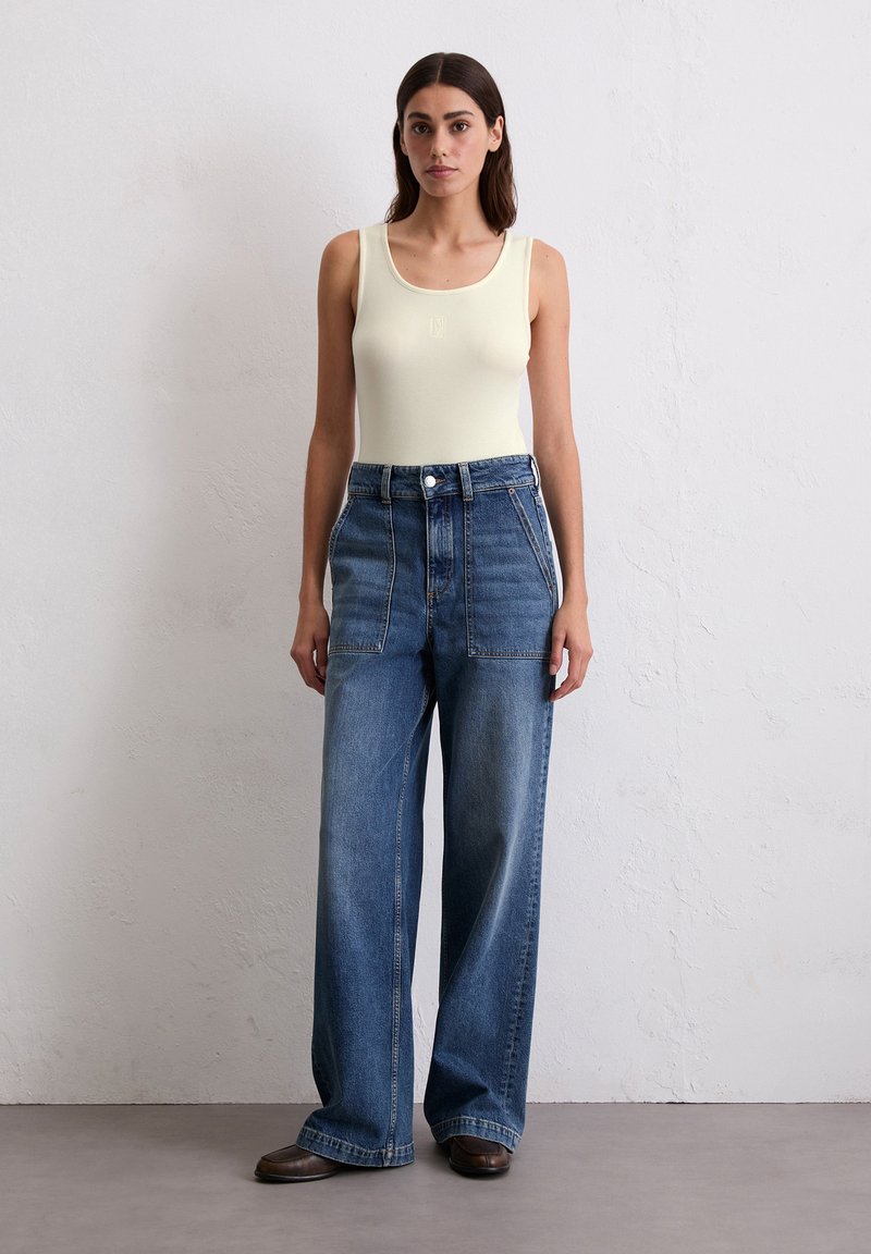 Kobieta stoi przy białej ścianie, ubrana w kremowy top na ramiączkach, spodnie dzwony w kolorze niebieskim z wysokim stanem i dużymi kieszeniami z przodu, oraz ciemne buty.