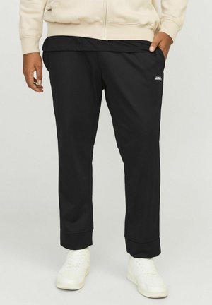Pantalon de survêtement noir en tissu doux, affichant une coupe ajustée, des poignets côtelés et un petit logo blanc sur la hanche gauche. Porté avec des baskets blanches.